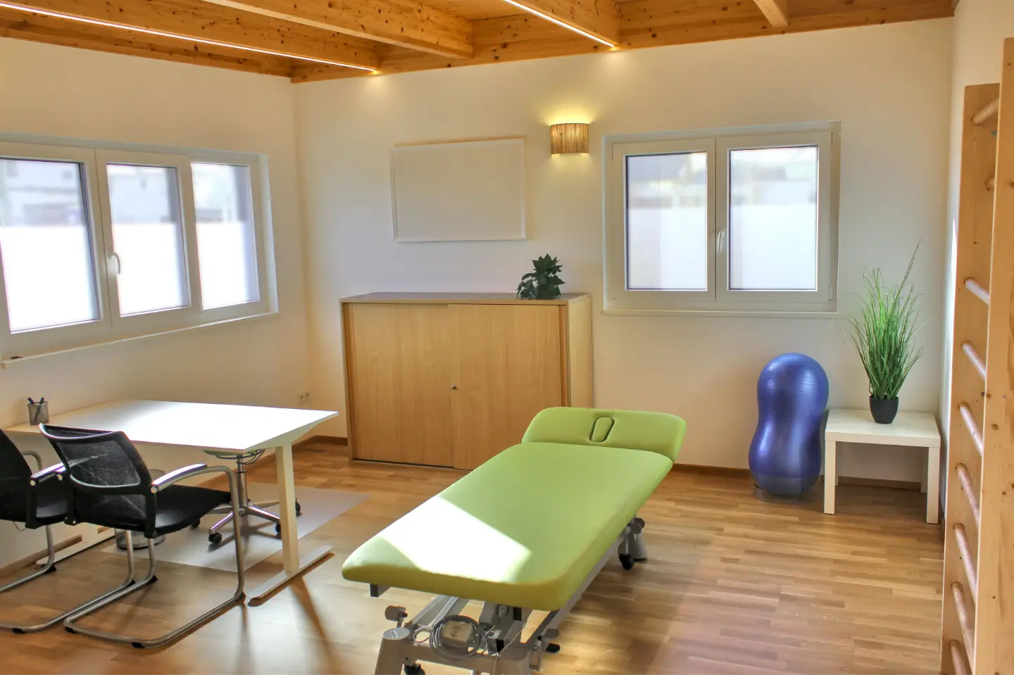 Therapieraum 1 in 2020 Sonnberg: Heller Raum mit Holzboden, in der Mitte eine grüne Liege, links davon ein weißer Schreibtisch mit zwei schwarzen Sesseln, in der rechten Ecke ein Tisch mit Pflanze und einem blauen Gymnastikball. An der rechten Wand ist eine Sprossenwand zu sehen. An der hinteren und linken Wand befinden sich Fenster.