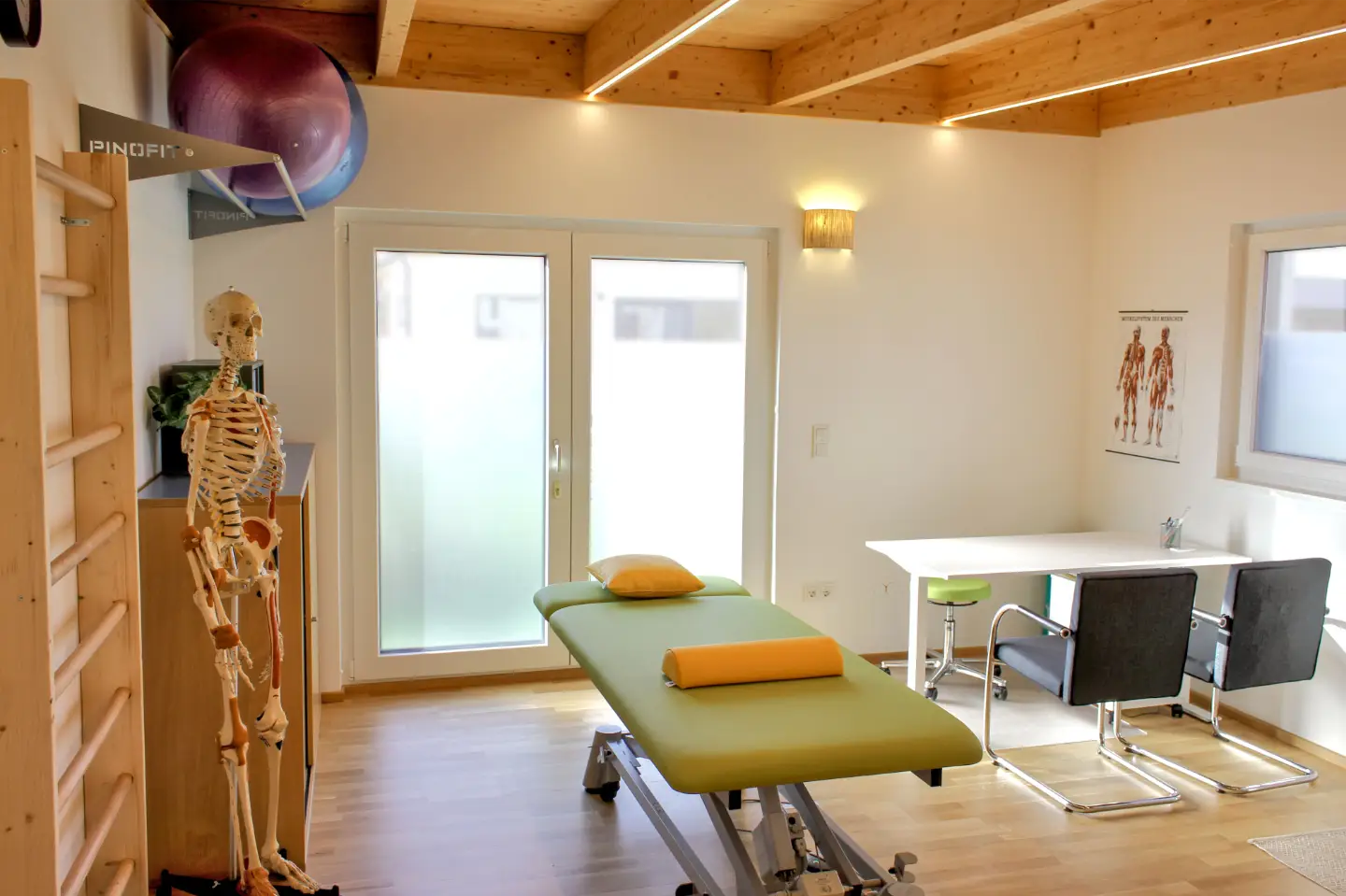 Therapieraum 2 in 2020 Sonnberg: Heller Raum mit Holzboden, in der Mitte eine grüne Liege mit zwei gelben Polstern, rechts davon ein weißer Schreibtisch mit zwei Sesseln, in der linken oberen Ecke eine Halterung mit zwei Gymnastikbällen (lila und blau). An der linken Wand ist eine Sprossenwand, davor steht ein Skelett. An der rechten Wand ist ein Fenster, an der hinterne Wand ein doppelflügige Terrassentür.